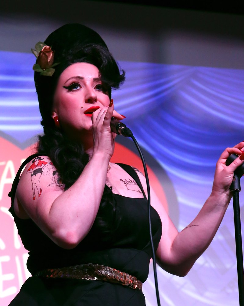 Natasha performing as Amy Winehouse, singing into the microphone on stage