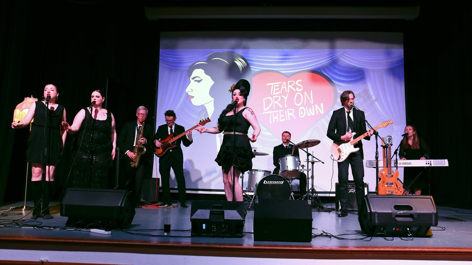 The full Tears Dry On Their Own band on stage in front of the Amy Winehouse show backdrop, with backing vocalists, saxophone, guitars, drums and keys