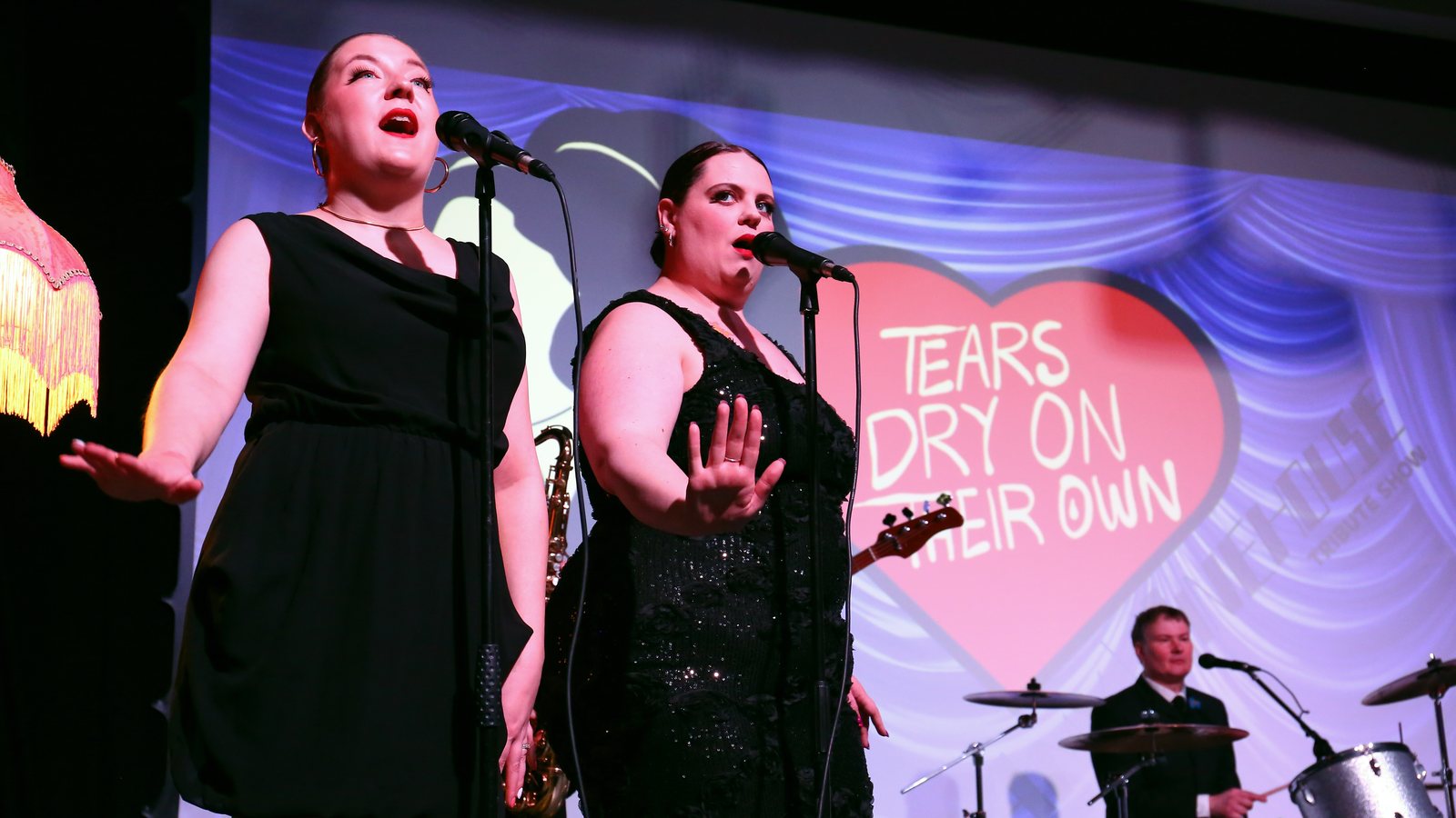 Emma and Melissa singing backing vocals beside the show backdrop, with saxophone and drums behind them